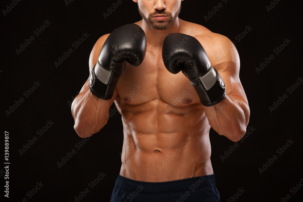 Muscular young man with perfect Torso with six pack abs, in boxing gloves is showing the different movements and strikes isolated on black background with copyspace
