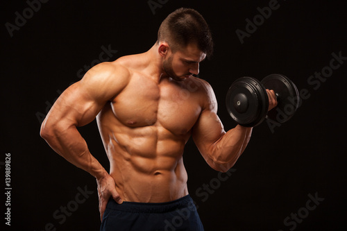 Handsome power athletic man with dumbbell confidently looking forward. Strong bodybuilder with six pack, perfect abs, shoulders, biceps, triceps and chest isolated on black background with copyspace