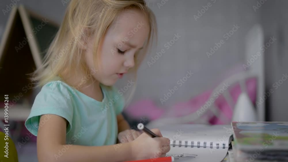 Little cute girl with blond hair, wearing a blue t-shirt sitting at the table and draws a colored felt-tip pens on a piece of paper. The girl focused on drawing, maybe she would be an artist Little cute girl with blond hair, wearing a blue t-shirt sitting at the table and draws a colored felt-tip pens on a piece of paper. The girl focused on drawing, maybe she would be an artist