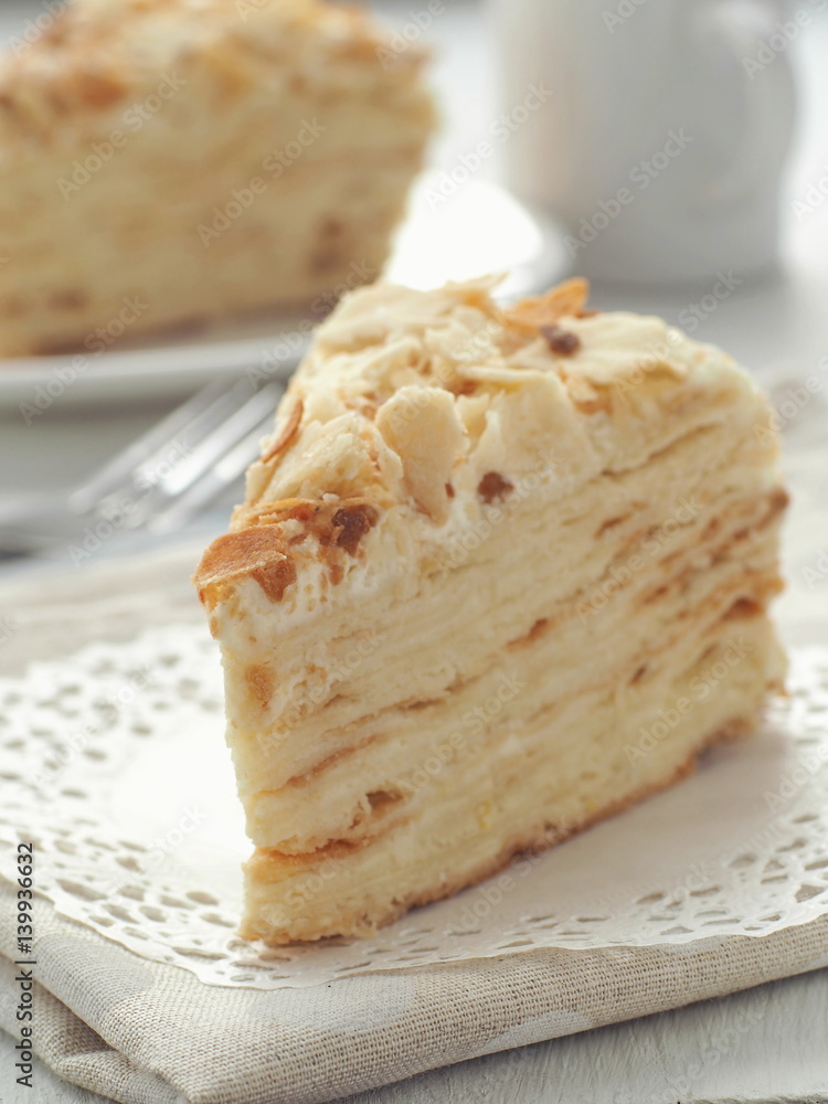 Piece of multi layered cake close-up. Mille feuille dessert. Crumbs decorated torte on white doily upon wooden table. Food background, blur. Selective focus on the front. Piece of multi layered cake close-up. Mille feuille dessert. Crumbs decorated torte on white doily upon wooden table. Food background, blur. Selective focus on the front.