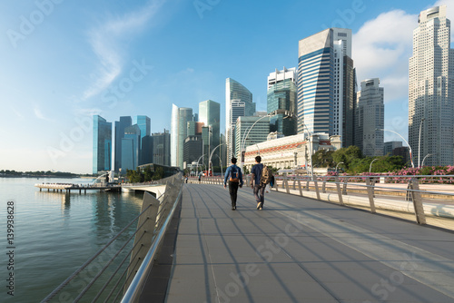 Quadro su tela Pedestrians walk along bridge near Marina bay in Singapore with Singapore skyscraper and Merlion park in background