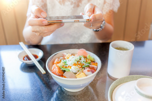Woman’s hands taking picture of Japanese sashimi dish by smartphone camera, soft-focus on hand and defocus in background.