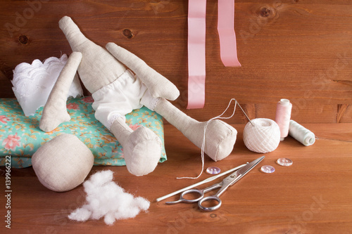 Textile doll in the sewing process with accessories and materials: lace, fabric, thread, needle, scissors. On wooden background photo