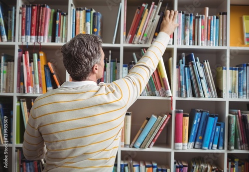Tablou pe pânză Rear view of school teacher selecting book in library