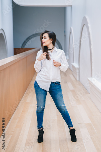 Portrait of smiling white Caucasian brunette young beautiful girl woman model with long dark hair in white shirt and blue jeans indoor in hall of college  university  standing full figure looking away