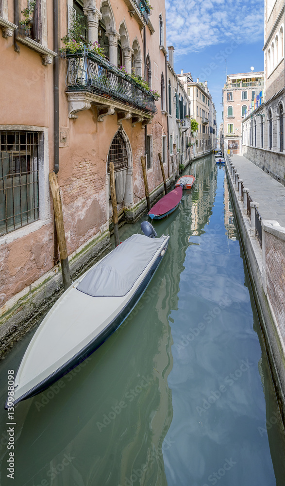 Narrow Venetian canal in the historic center with row of boats moored and covered next to the typical stone buildings, Venice, Italy