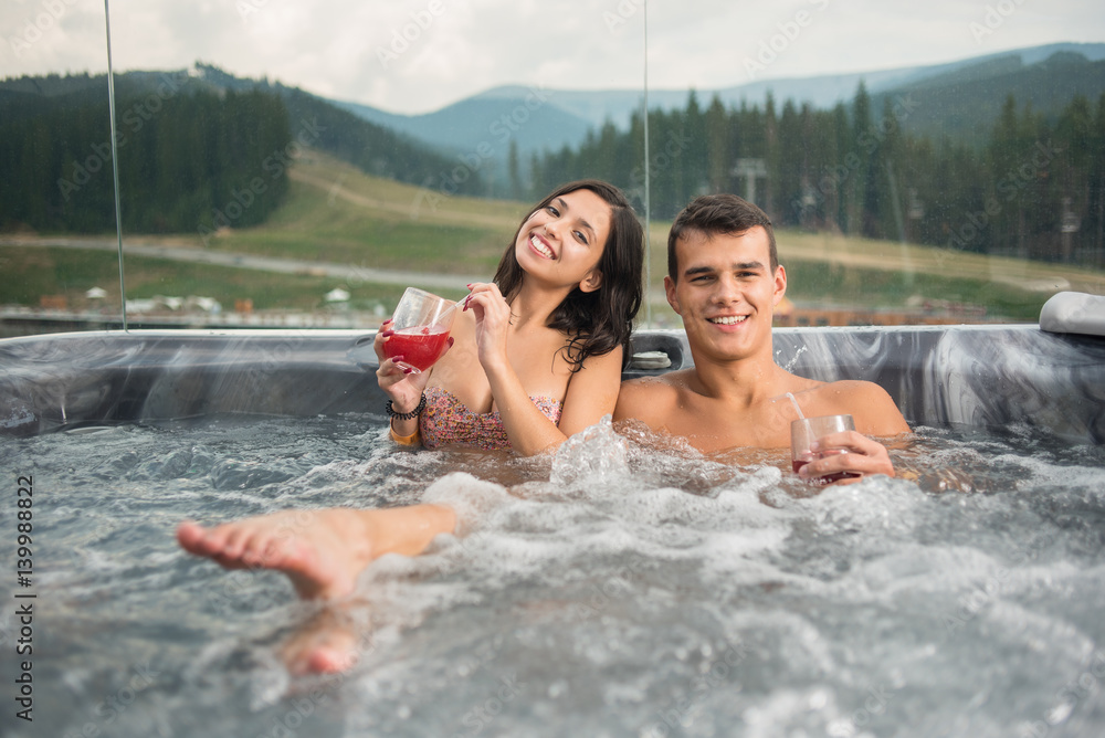 Happy young couple enjoying a bath in Jacuzzi while drinking cocktail outdoors on romantic vacation against blurred background of nature - obrazy, fototapety, plakaty 