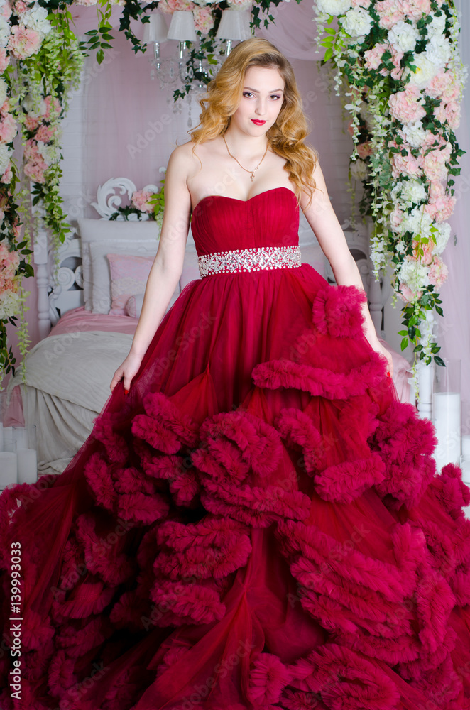 Charming woman in a beautiful lush dress cloud royal burgundy. She stands by the bed, decorated with flowers Charming woman in a beautiful lush dress cloud royal burgundy. She stands by the bed, decorated with flowers