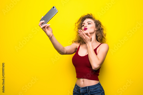 Young beautifulgirl with an curly hairstyle send selfie kiss. Laughing girl take selfie from phone on Yellow background. photo