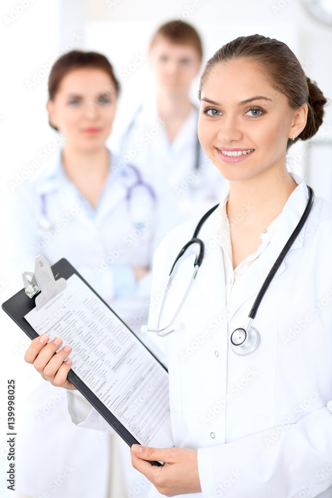 Happy female doctor keeping medical clipboard while medical staff are at the background. Successful team at health care and medicine concept Happy female doctor keeping medical clipboard while medical staff are at the background. Successful team at health care and medicine concept