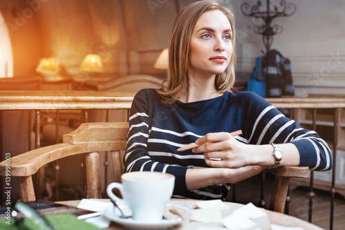 A smile young woman dreams seat in a cafe with cup of coffee with wooden pencil in hand. Concept of planning personal traning schedule. Looking away.