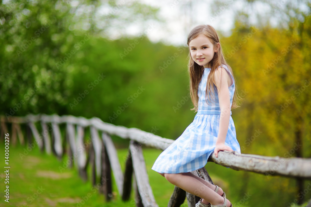 Fototapeta premium Adorable little girl sitting on a wooden fence on beautiful spring day