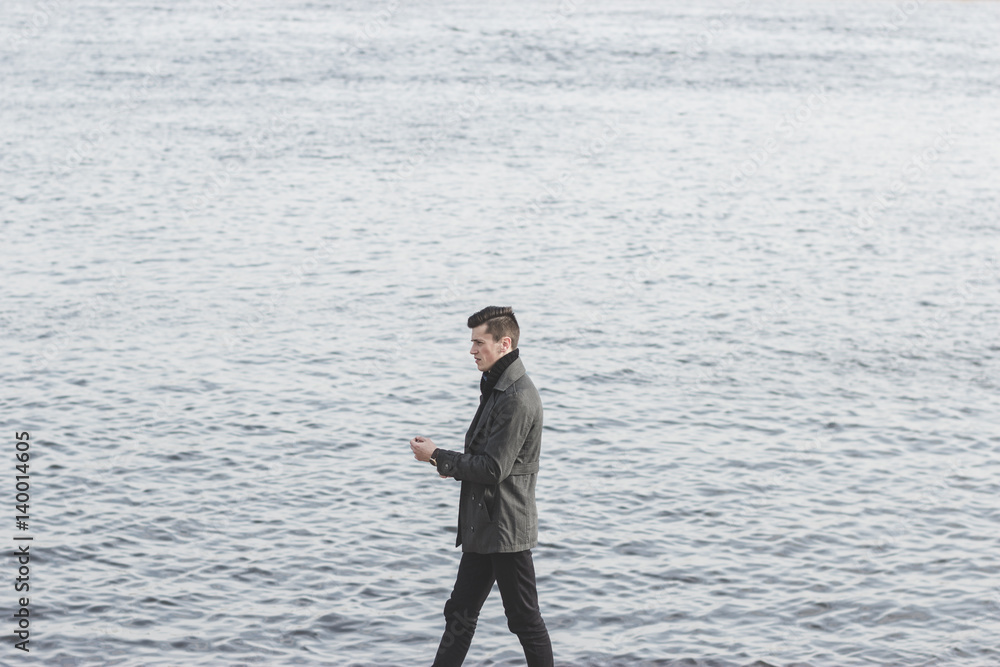 handsome young man in coat walking near water handsome young man in coat walking near water