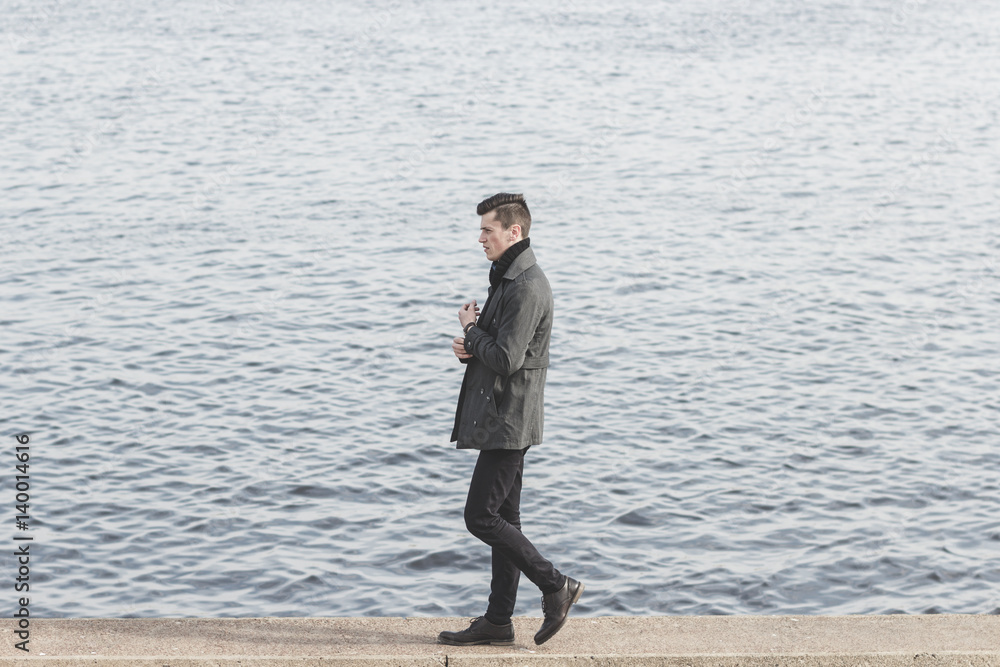 handsome young man in coat walking near water handsome young man in coat walking near water