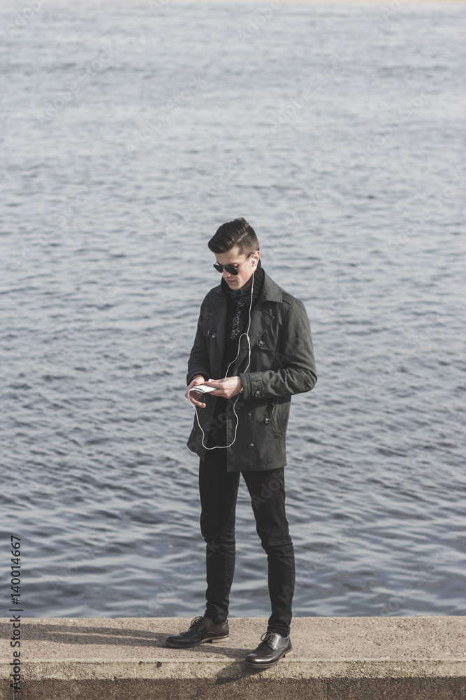 handsome young man in sunglasses listening to music near water