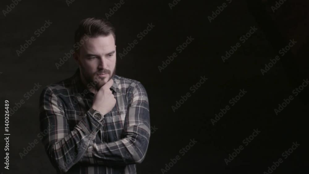 Bearded young man posing in the studio on a black background. A man with a beard with his hand touching his chin Bearded young man posing in the studio on a black background. A man with a beard with his hand touching his chin