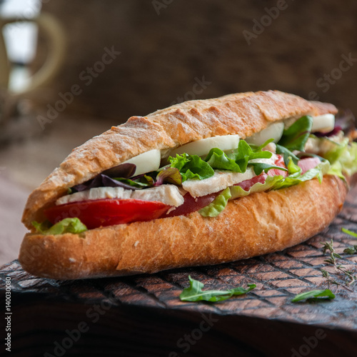 Long baguette sandwich with lettuce tomatoes eggs onion and mayo sauce served on a wooden board. Rustic style and close up. Square image.
