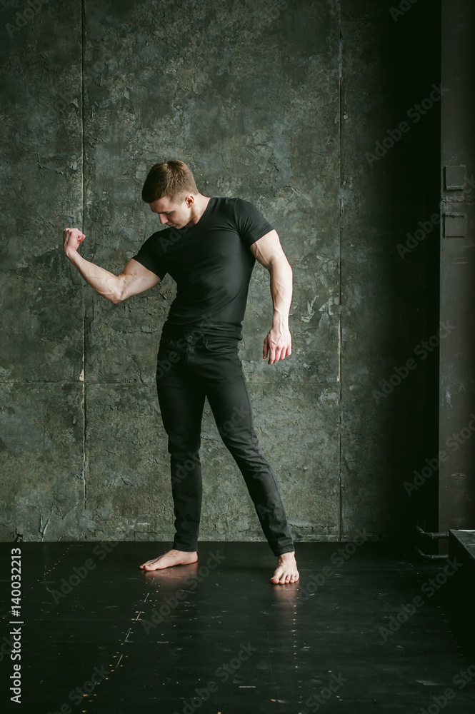 Fototapeta premium Young sexy men bodybuilder athlete, studio portrait loft on background of stylized Concrete brutal wall, guy model black Tshirt and trousers With powerful muscular arms demonstrates strength