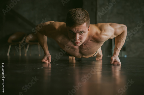 Young sexy male bodybuilder athlete with a bare torso. portrait of a studio of a light-skinned strong man engaged in push-ups on the fists, on the floor, straining powerful hands
