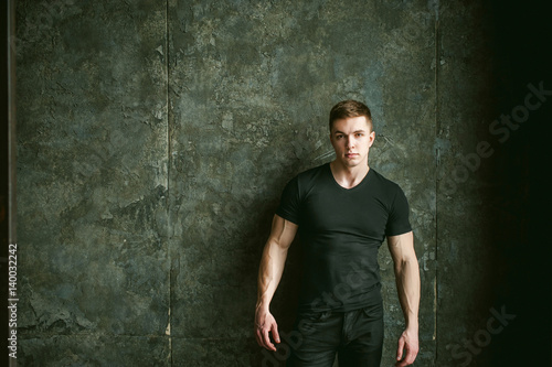 Young sexy men bodybuilder athlete, studio portrait loft on background of stylized Concrete brutal wall, guy model black Tshirt and trousers With powerful muscular arms