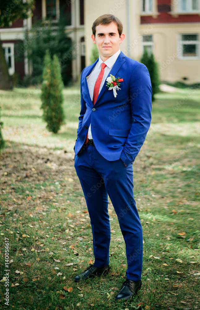 Handsome groom in bright blue suit poses on green lawn