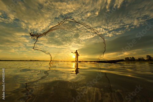 Thai fisherman on wooden boat casting a net for catching freshwater fish in nature river in the early evening before sunset photo