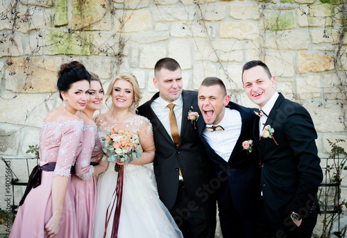 Funny groomsmen, bridsmaids and newlyweds pose before white stonewall photo