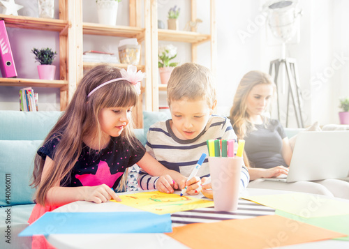 Siblings drawing with colorful pencils together while mother working on laptop