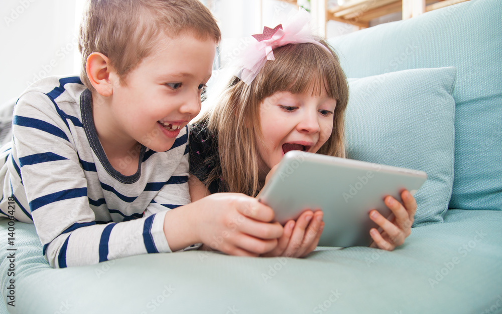 Happy siblings lying on sofa at home and playing with tablet together Happy siblings lying on sofa at home and playing with tablet together