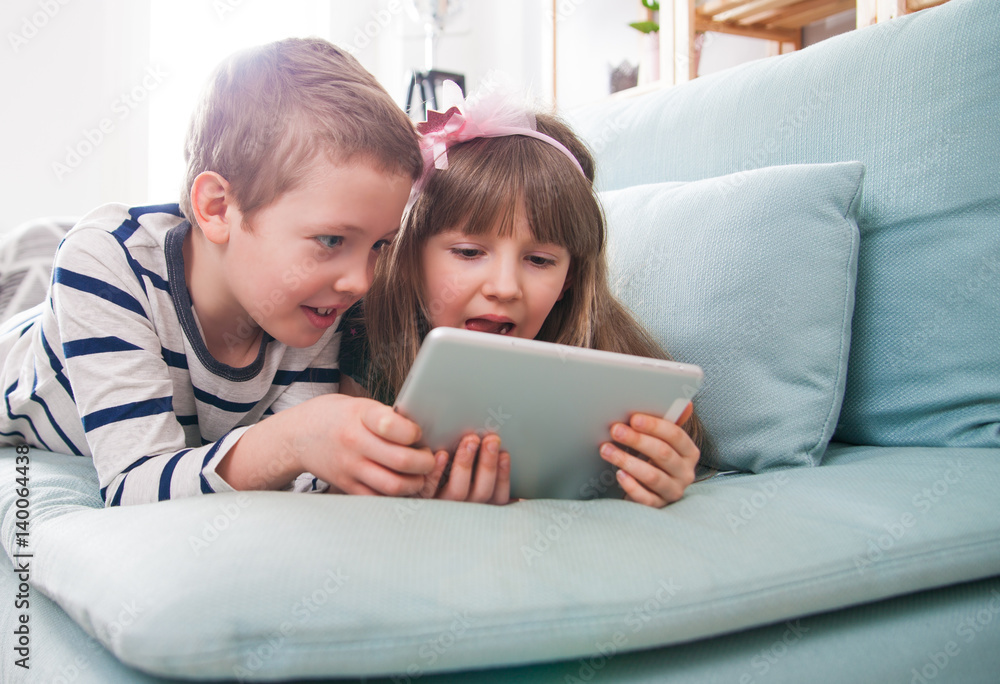 Happy siblings lying on sofa at home and playing with tablet together Happy siblings lying on sofa at home and playing with tablet together