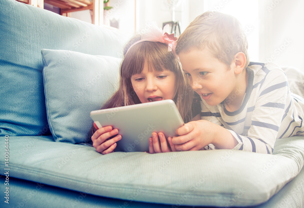 Happy siblings lying on sofa at home and playing with tablet together Happy siblings lying on sofa at home and playing with tablet together