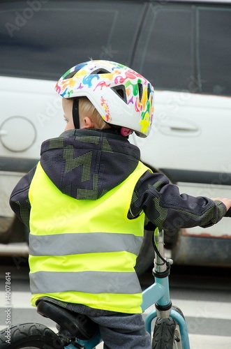 Danger situation in the city traffic. Little child rides a bike across the zebra crossing while white car is driving in front of him. Boy is marked by yellow reflective vest and helmet.