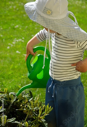 Little boy is watering plants in the garden wit his green can. He has has as a sun protection.