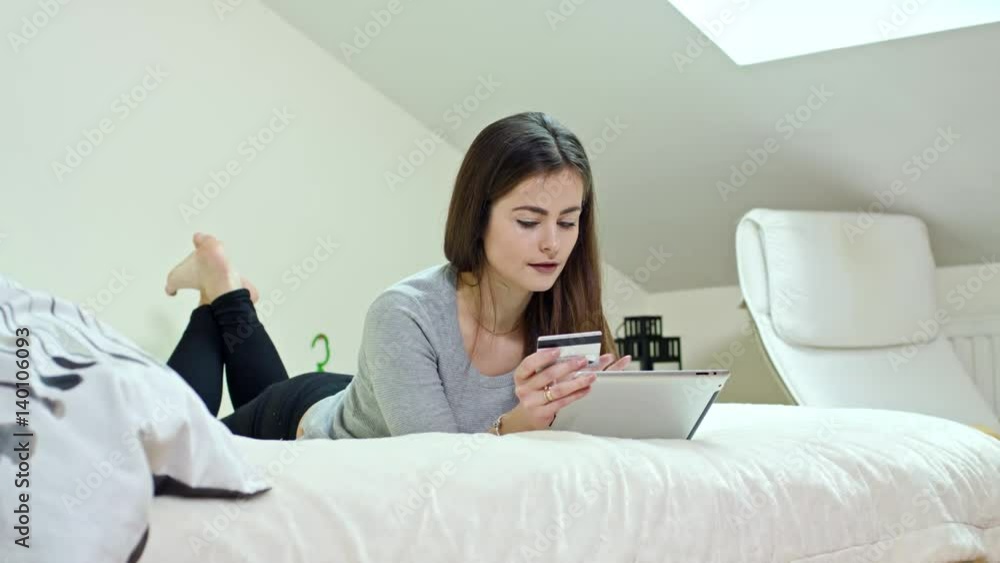 Smiling nice young woman uses a digital tablet and credit card lying on a bed at home, on-line shopping Smiling nice young woman uses a digital tablet and credit card lying on a bed at home, on-line shopping