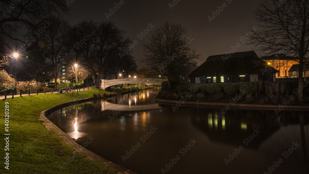 A night time photograph of a river running through a town park with a bridge in the distance and reflections in the water