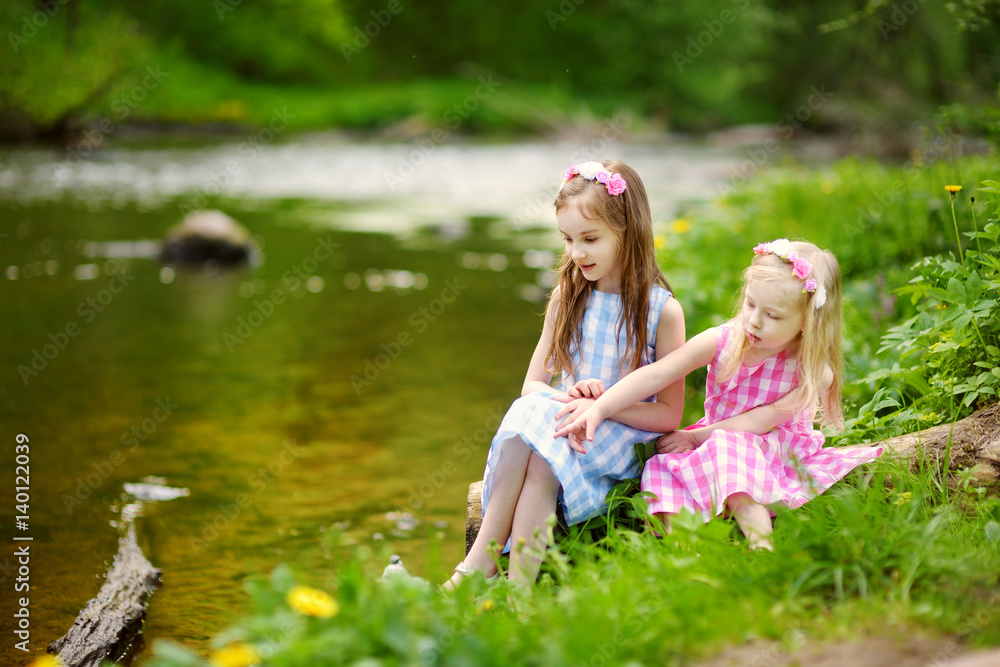 Two adorable little sisters playing by a river in sunny park on a beautiful summer day Two adorable little sisters playing by a river in sunny park on a beautiful summer day
