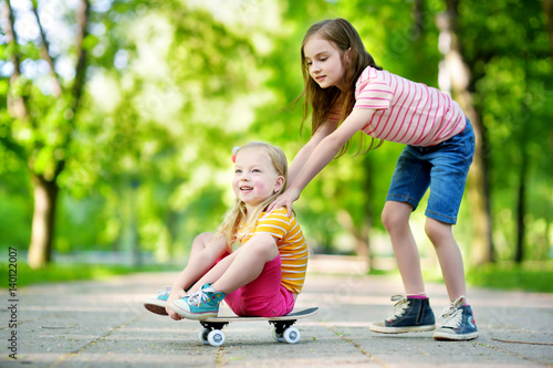 Two pretty little girls learning to skateboard on beautiful summer day in a park