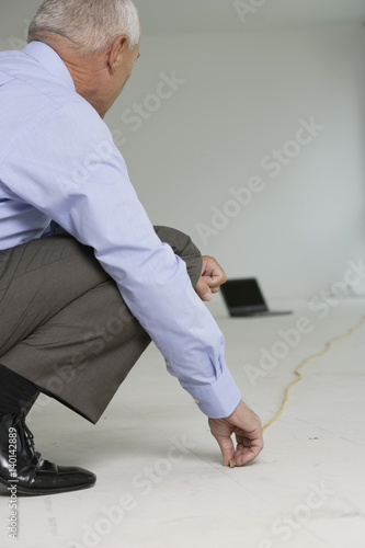 Elderly manager with folding rule squatting on the floor, laptop on floor in the background