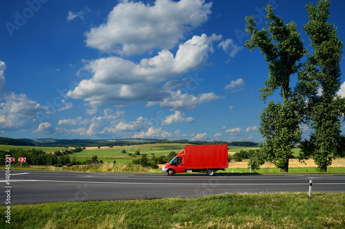Red delivery truck on asphalt road in a rural landscape under a blue sky with dramatic clouds