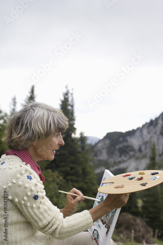 Senior adult woman sitting on a meadow while painting with a color palette, close-up © Gudrun