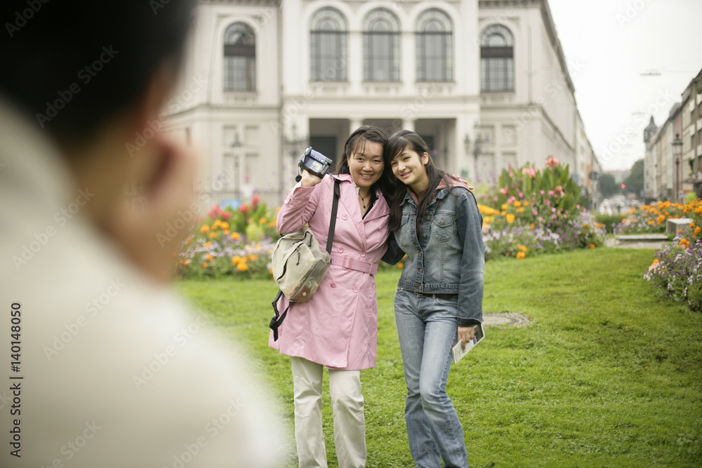 Man taking a picture of two Asian women who are standing on a meadow in front of buildings, selective focus