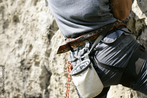 Young man with a harness and a chalk bag is climbing up a rocky wall (part of), close-up