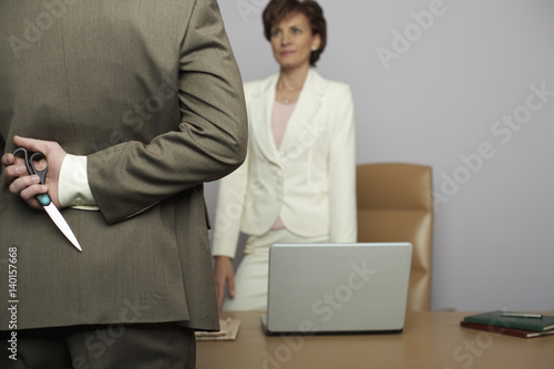 Businessman holding scissors behind back, facing businesswoman in office