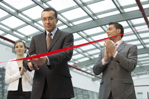 Businessman cutting red ribbon, colleagues clapping