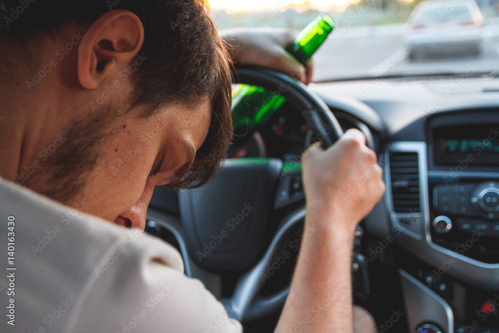 Drunk young man driving a car with a bottle of beer. Don't drink and drive concept. Driving under the influence. DUI, Driving while intoxicated. DWI Drunk young man driving a car with a bottle of beer. Don't drink and drive concept. Driving under the influence. DUI, Driving while intoxicated. DWI