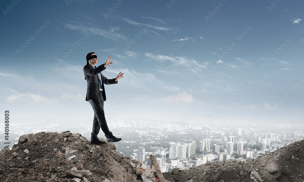 Young businessman in blindfold walking carefully and cityscape at background Young businessman in blindfold walking carefully and cityscape at background