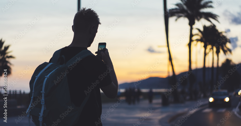 Fototapeta premium Man with backpack making photo sunset beach on smart phone, tourist look and enjoy nature on background color sky and taxi on palm summer street, hipster using in hands mobile phone, traveler mock up