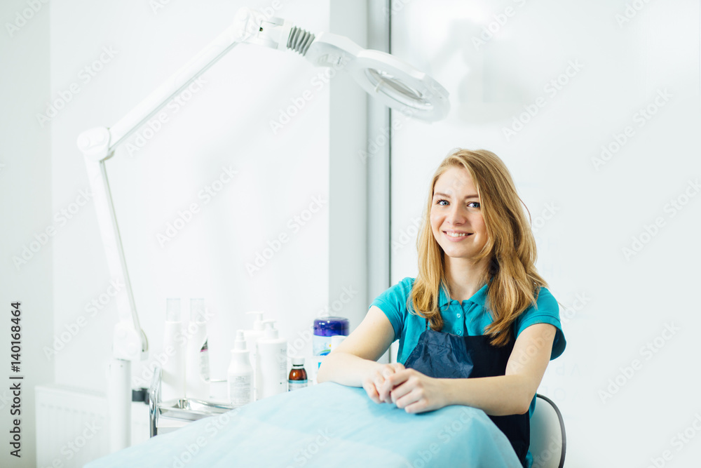Portrait of young female beautician in turquoise t-shirt waiting for client in workplace of cosmetology clinic. Portrait of young female beautician in turquoise t-shirt waiting for client in workplace of cosmetology clinic.