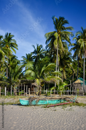 Depth of field shot of a basic Filipino pump boat on the shore of a white sand beach with a back ground of fenced area, palm trees and a basic bamboo hut.
