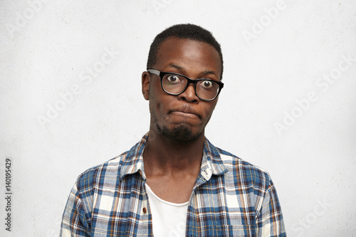 Funny emotional bug-eyed dark-skinned male student wearing spectacles holding breath with pursed lips, blowing his cheeks, having frustrated look, not supposed to say anything. Body language